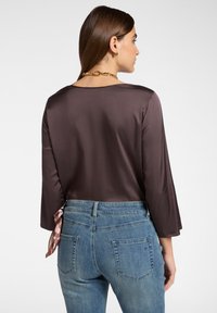 Zijdeachtige bruine top met lange mouwen, brede halslijn en strikdetail aan de achterkant, gecombineerd met blauwe denim jeans met achterzakken.