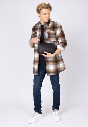 Rutig flanellskjorta i brunt, vitt och orange. Slim-fit jeans och vita sneakers. Håller en svart läder rektangulär väska med logotyp.