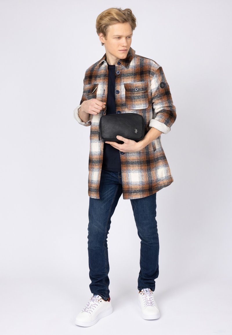 Rutig flanellskjorta i brunt, vitt och orange. Slim-fit jeans och vita sneakers. Håller en svart läder rektangulär väska med logotyp.