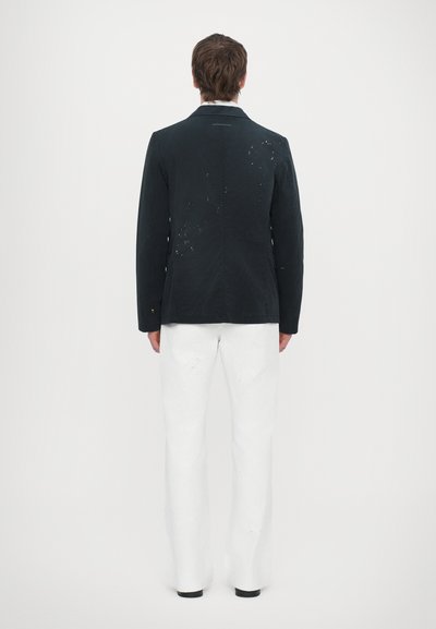 Chaqueta negra con detalles texturizados y manchas de pintura blanca, combinada con pantalones anchos blancos. Presentada desde la parte trasera sobre un fondo neutro.