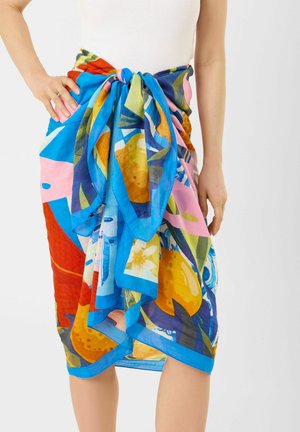 Heldere, meerkleurige sjaal met een bloemig en fruitig patroon, gestrikt als een knie-lange rok bij een persoon die een witte mouwloze top draagt.