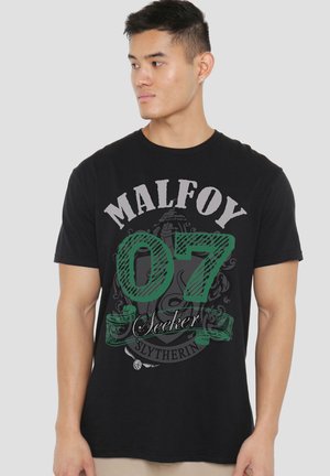Junger Mann trägt schwarzes T-Shirt mit "Malfoy 07 Seeker Slytherin" Harry Potter-Design und blickt nach links vor einfarbigem Hintergrund.
