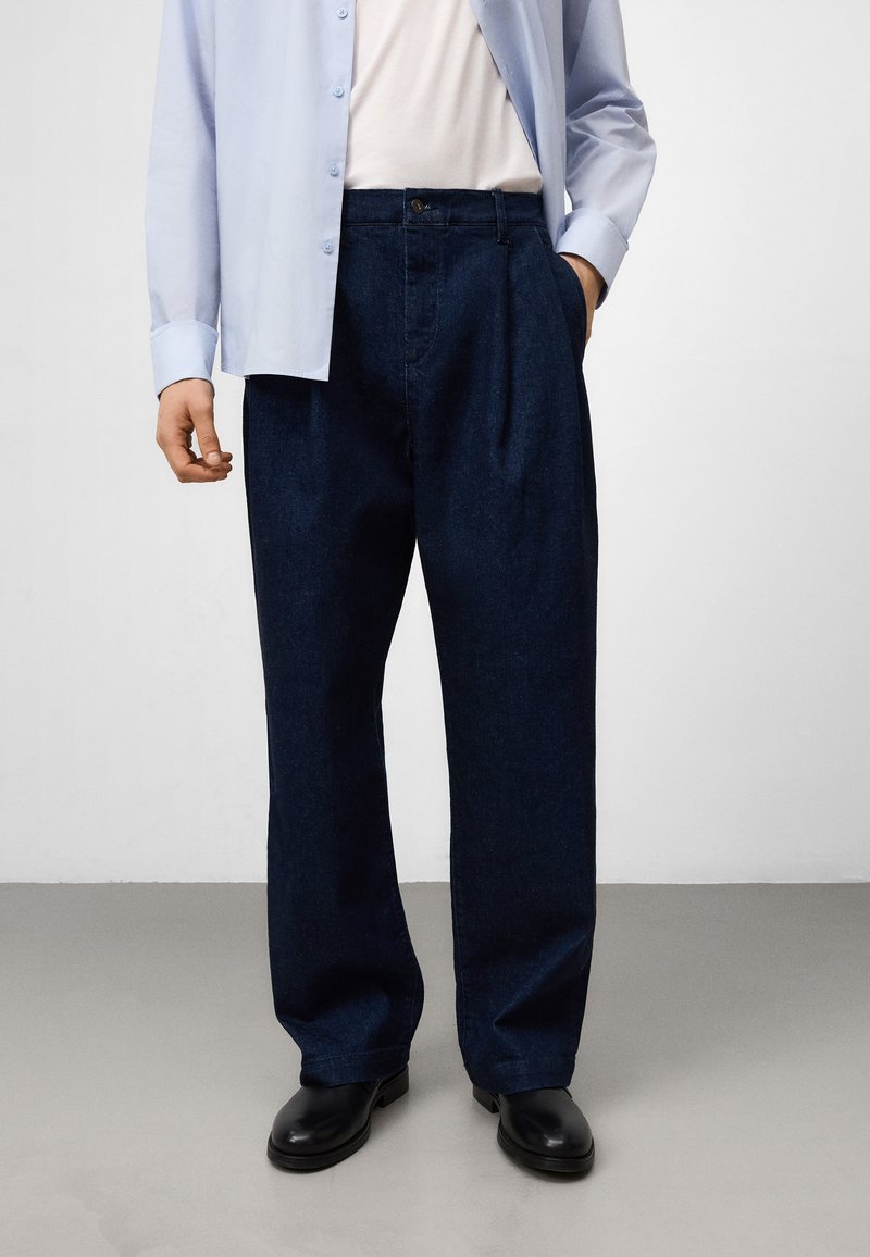Mann in dunkelblauen, weit geschnittenen Jeans, schwarzen Schuhen, weißem T-Shirt und hellblauer, kurzärmelliger Hemd steht auf grauem Boden.