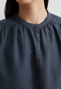 Donkerblauwe blouse met knopen en een ronde halslijn, met geplooide details op de schouders en een gladde textuur. Bevat kleine knopen aan de voorkant.