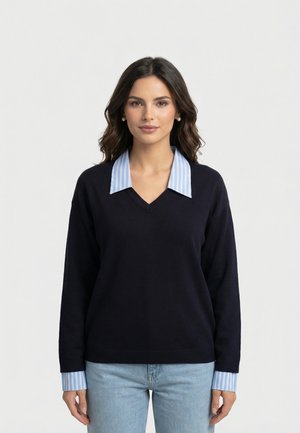 Donna con capelli scuri e mossi indossa un maglione a V nero sopra una camicia a righe blu e bianche con colletto e jeans azzurri, in piedi su uno sfondo bianco.