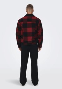 Jachetă din flanel cu carouri roșii și negre, cu guler, manșete cu nasturi și buzunar pe piept. Asortată cu pantaloni negri și încălțăminte casual.