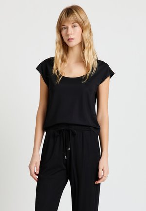 Giovane donna con lunghi capelli biondi che indossa una maglietta nera a maniche corte e pantaloni neri con coulisse, su uno sfondo chiaro e uniforme.