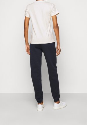 Persona de pie de espaldas, vistiendo una camiseta blanca de manga corta lisa, pantalones deportivos negros y zapatillas blancas sobre un fondo blanco.