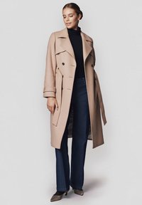 Beżowy wełniany płaszcz trench z podwójnym zapięciem na guziki, pasek w talii i szerokimi klapami, w zestawieniu z granatowym golfem i dzwonami.