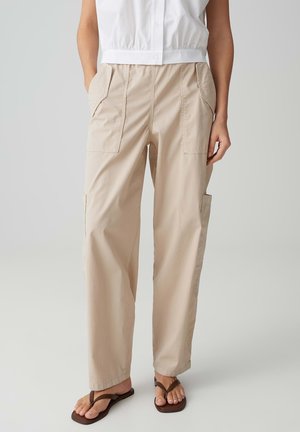 Pantalon beige à jambes larges avec grandes poches avant et latérales, porté avec une chemise boutonnée blanche sans manches et des tongs marron sur un fond neutre.