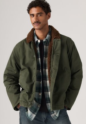 Homme portant une veste verte avec un col en polaire marron par-dessus une chemise à carreaux noire et verte et un jean bleu devant un fond uni.