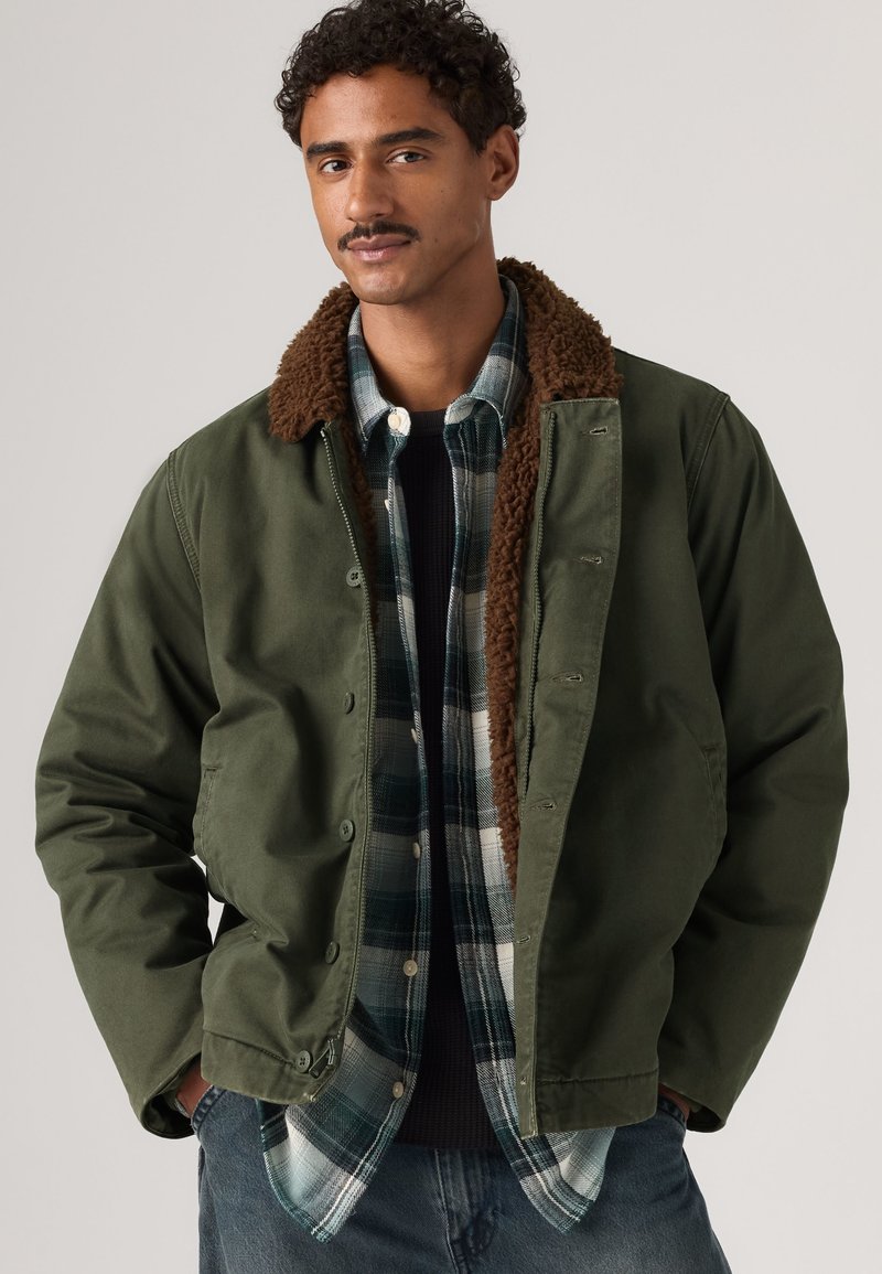 Homme portant une veste verte avec un col en polaire marron par-dessus une chemise à carreaux noire et verte et un jean bleu devant un fond uni.