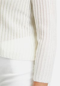 Bílý pletený svetr s vertikálním žebrováním, texturovanou tkaninou a tříčtvrtečními rukávy. Spodní lem končí těsně nad pasem.