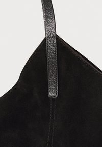 Sac en daim noir avec une sangle en cuir texturé, des angles marqués et des coutures minimales. Le design général est élégant et moderne.