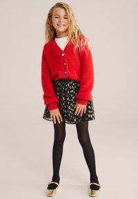 Piros bolyhos kardigán fekete gombokkal, fekete szoknyával, amely arany- és fehér mintával díszített, fekete harisnyával és arany balettcipőkkel párosítva.