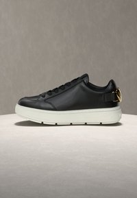 Μαύρο δερμάτινο αθλητικό παπούτσι με λευκή χοντρή σόλα, μαύρα κορδόνια και χρυσό μοτίβο σε σχήμα καρδιάς στο λουρί της φτέρνας, τοποθετημένο σε ανοιχτή επιφάνεια.