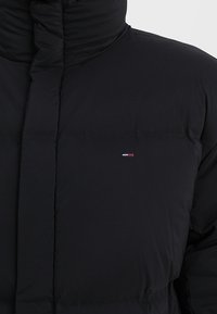 Giacca imbottita nera con collo alto, texture trapuntata e piccola toppa con logo sul petto sinistro. Materiale morbido e isolante.