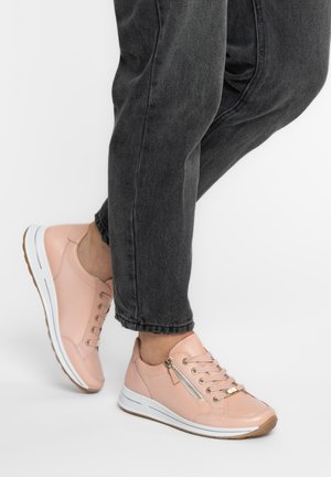 Person trägt hellrosa Leder-Sneaker mit seitlichen Reißverschlüssen und weißen Sohlen, kombiniert mit schwarzer Jeans, stehend auf einer weißen Oberfläche.
