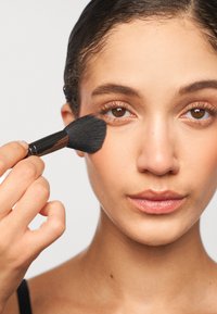 Black makeup brush held near cheek, applying product. Model has smooth skin, light makeup, brown eyes, and dark hair pulled back.