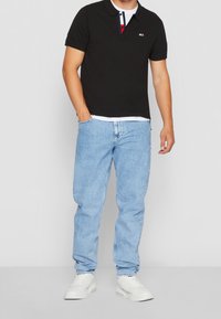 Svart polo skjorta med krage och kontrasterande knappar, i kombination med ljusblå jeans och vita sneakers. Lätt struktur synlig på skjortan.