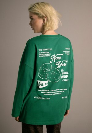 Green long-sleeve t-shirt featuring white text and donut graphics, with a relaxed fit and rounded neckline. Text includes shop details and prices.