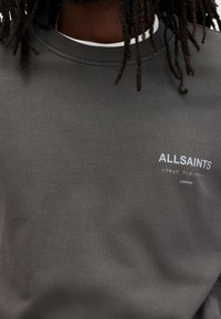 Grå sweatshirt med rund hals, tillverkad av bomull. Har en vit "ALLSAINTS" logotyp och text på både engelska och japanska.