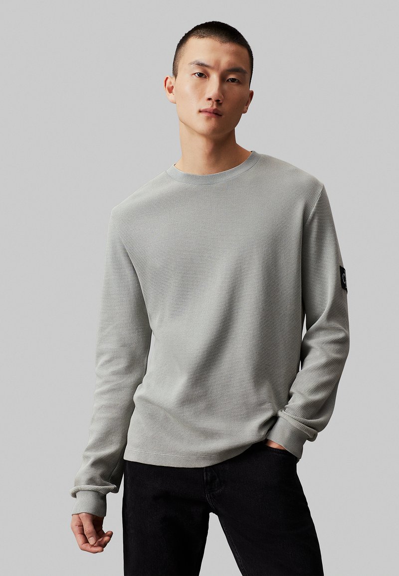 Camisa de manga larga gris claro con textura acanalada, cuello redondo y parche de logo negro en la manga izquierda, combinada con jeans oscuros.