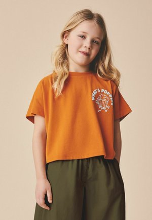 Noor tüdruk pikkade blondide juustega, kes kannab oranži lühikest T-särki kirjaga "What's Poppin" ja popkorni graafikuga, kombineeritud oliivroheliste pükstega.