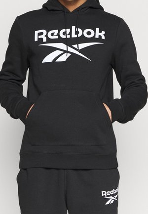 Personne portant un sweat à capuche noir Reebok et un pantalon de survêtement noir assorti avec des logos blancs Reebok sur la poitrine et la partie supérieure de la cuisse gauche.