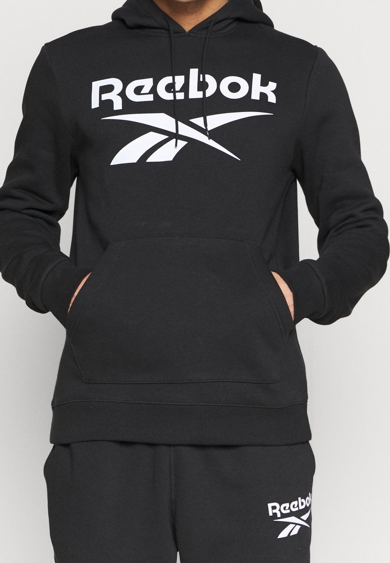 Personne portant un sweat à capuche noir Reebok et un pantalon de survêtement noir assorti avec des logos blancs Reebok sur la poitrine et la partie supérieure de la cuisse gauche.