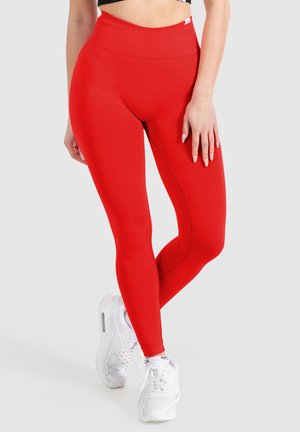 Leggings rouges taille haute en tissu extensible. Texture lisse avec un ajustement serré, associés à des chaussures de sport blanches.