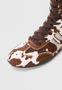 Schuh mit braun-weißem Kuhfellmuster, runder Spitze, Schnürverschluss und strukturierter Oberfläche. Leicht, mit schwarzer Gummisohle.