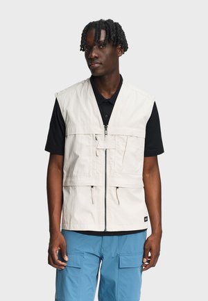 Muškarac u bež prsluku bez rukava preko crne majice kratkih rukava i plavih cargo kratkih hlača, stoji ispred jednobojne pozadine.