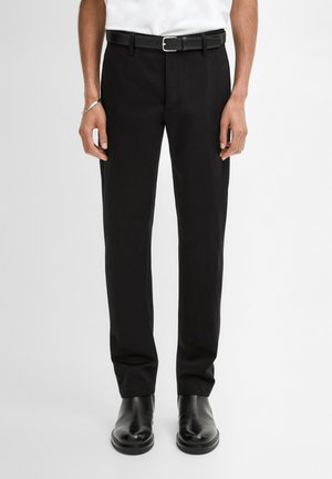 Homme portant un pantalon habillé noir avec une ceinture noire et des chaussures en cuir noires, debout devant un fond blanc uni.