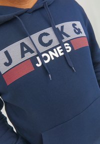 Sudadera azul marino con texto gráfico en rayas blancas y rojas que dice "JACK & JONES". Presenta un bolsillo tipo canguro en la parte frontal y cordones en la capucha.