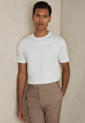 T-shirt blanc à manches courtes en tissu lisse, col rond, associé à un pantalon texturé marron. Arrière-plan dans des tons neutres.