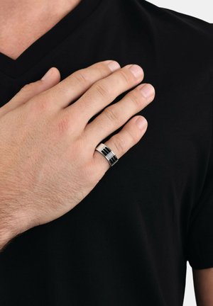 Silver ring with black accents, featuring a smooth surface and geometric patterns, worn on a hand against a black v-neck shirt.