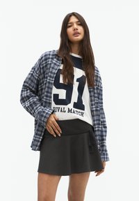 Chemise en flanelle bleu à carreaux, t-shirt graphique blanc avec "51 RIVAL MATCH," et jupe évasée noire avec un détail de lien à la taille.