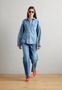 Camisa y pantalones de mezclilla azul claro en conjunto, con dos bolsillos en el pecho. Combinado con zapatos naranjas y gafas de sol marrones. Diseño simple.