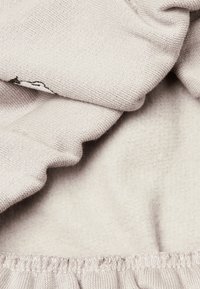 Tessuto beige chiaro con una texture morbida, caratterizzato da bordi arricciati e un semplice design ricamato. Il materiale appare accogliente e caldo.
