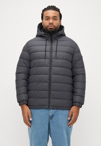 Γκρι μπουφάν τύπου φουσκωτό με κουκούλα, με οριζόντια σχεδίαση καπιτονέ, ρυθμιζόμενα κορδόνια και ελαστικά μανίκια, φορεμένο πάνω από ανοιχτόχρωμα μπλε τζιν.