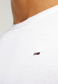 Detailní záběr na osobu oblečenou v bílém tričku s kulatým výstřihem, na hrudi má malé vyšívané logo v červeno-bílo-modrých barvách v obdélníkovém tvaru.