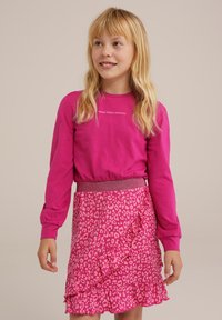 Dívka s blond vlasy, oblečená v růžovém tričku s dlouhým rukávem, na kterém je nápis "Make Today Amazing", a v růžové sukně s leopardím vzorem a volánky.