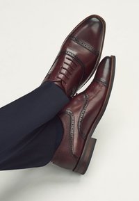 Chaussures habillées en cuir bordeaux foncé avec détails brogue, bout rapporté et coutures élaborées, associées à un pantalon texturé bleu marine.