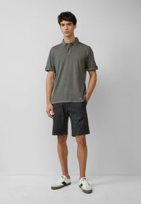Grijze polo met opgerolde mouwen, gecombineerd met donkere shorts. Model draagt witte sneakers met bruine accenten en staat tegen een neutrale achtergrond.