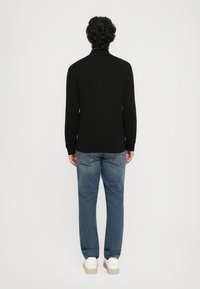 Mann mit lockigem Haar, der ein schwarzes Langarmoberteil, blaue Jeans und weiße Sneaker trägt und mit dem Rücken zu einem schlichten Hintergrund steht.