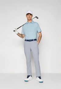 Svetlomodrá polokošeľa so vzorom, sivé nohavice, biele golfové topánky s modrými doplnkami. Drží golfovú palicu, má na sebe bielu čiapku a golfovú rukavicu.