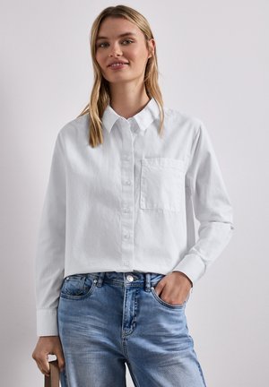 Kobieta w białej koszuli na guziki z kieszenią na piersi i niebieskich dżinsach, stojąca z jedną ręką w kieszeni i lekkim uśmiechem.