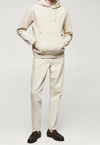 Homme portant un sweat à capuche blanc cassé, un pantalon assorti de la même couleur et des mocassins en cuir brun foncé, debout les mains dans les poches.