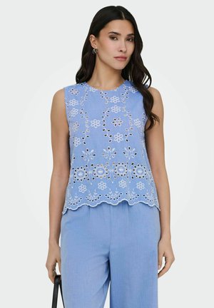 Femme portant un haut sans manches bleu clair brodé avec découpe florale et un pantalon assorti, tenant un sac à main noir.